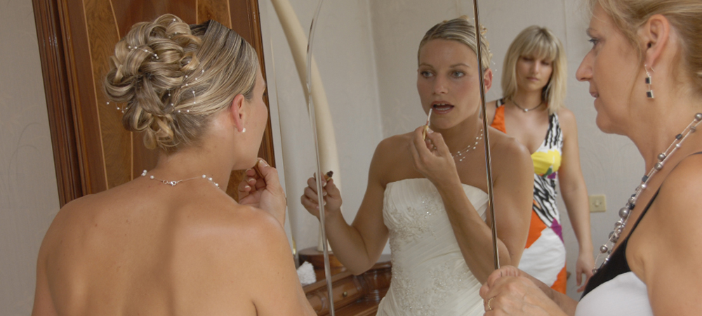 Mariée en robe blanche ajustant son maquillage devant un miroir, entourée de ses proches, préparation de mariage, coiffure élégante avec bijoux scintillants, moment émotionnel et complicité avant la cérémonie nuptiale.