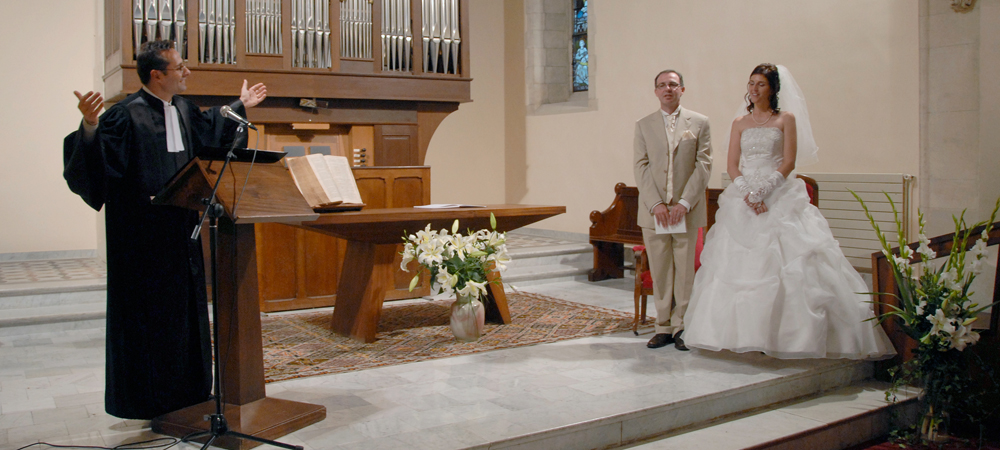 Cérémonie de mariage protestant dans une église, marié et mariée écoutant le pasteur en robe noire, ambiance solennelle avec orgue en arrière-plan, bouquets de fleurs blanches et décoration sobre, moment sacré et émouvant.