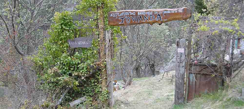 Panneau en bois indiquant 'Exploitation agricole - Towanda' au début d'un sentier, entouré de végétation sauvage et d'arbres, entrée rustique. 