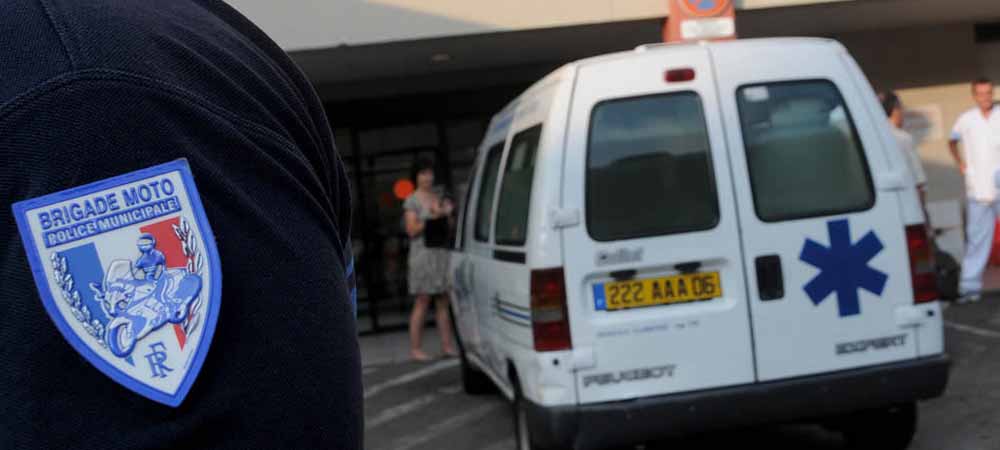 Uniforme de la brigade moto de la police municipale avec écusson visible, véhicule médicalisé blanc marqué d'une croix bleue garé devant un bâtiment, scène d'intervention ou de secours en milieu urbain.