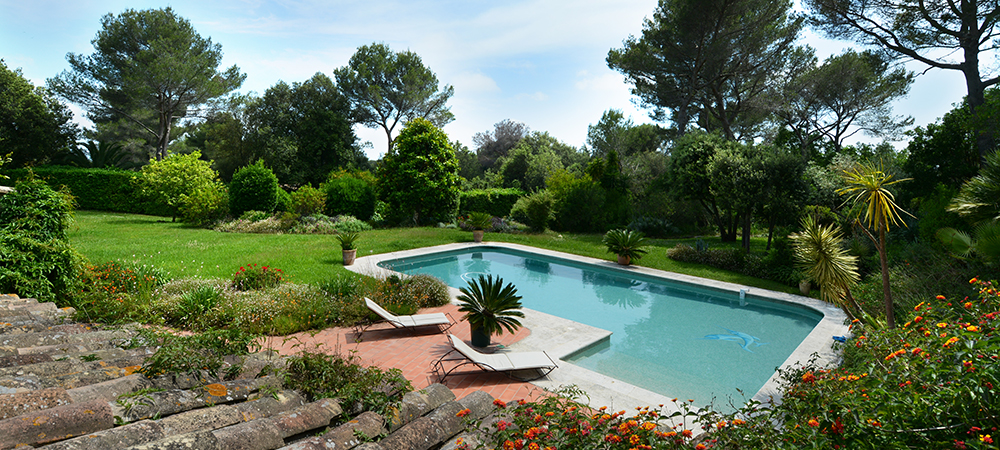 Piscine extérieure entourée d'un jardin méditerranéen luxuriant, transats en teck disposés sur une terrasse en pierres naturelles, vue dégagée sur une végétation dense et variée, propriété haut de gamme avec espace détente et intimité garantie.